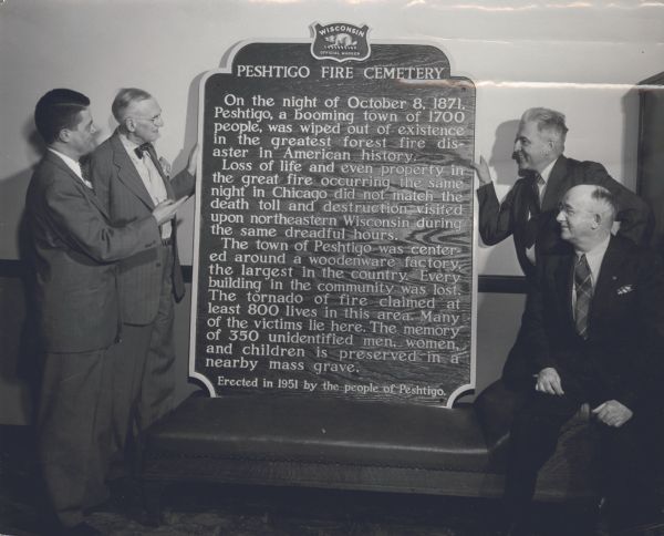 Indoor group portrait of men posed with the Wisconsin Official Marker. From left to right on either side of the marker are: Clifford Lord, Secretary of Sites and Markers Committee and Director of State Historical Society; James Law, Chairman of Highway Dept and Chairman of Sites and Markers Committee; C.L. Harrington, Superintendent of Forests and Parks, Wisconsin Conservation Dept; Russell Williams, Roadside Development Engineer, State Highway Commission.<p>The text on the Peshtigo Fire Cemetery marker reads:<p>"On the night of October 8, 1871, Peshtigo, a booming town of 1700 people, was wiped out of existence in the greatest forest fire disaster in American history.<p>Loss of life and even property in the great fire occurring the same night in Chicago did not match the death toll and destruction visited upon northeastern Wisconsin during the same dreadful hours.<p>The town of Peshtigo was centered around a woodenware factory, the largest in the country. Every building in the community was lost. The tornado of fire claimed at least 800 lives in this area. Many of the victims lie here. The memory of 350 unidentified men, women, and children is preserved in a nearby mass grave."