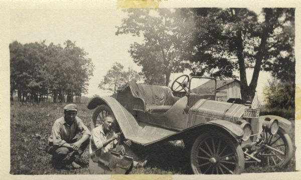 Two men sitting on the ground near a 1914-1916 touring car. A small barn is in the background behind the car. The photograph is annotated on the back:<p>"Latest thing in transportation - 2 good fire fighters."