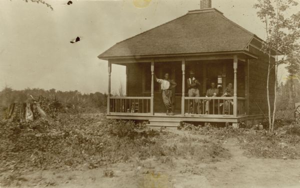 Four men are lounging on the porch of a small house in the middle of a cleared forest plot. Two of the men are holding dogs. To the right of the house is the wheel of a wagon.