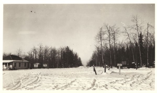 View across snow towards a concession building on the left and a parked truck on the right. Caption on back reads: "Concession building is at the left. The open cut through the woods is an old unused road which can be developed into a future parking area."