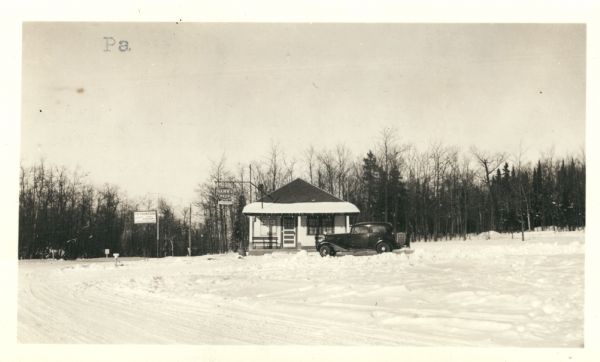 View across snow towards concession building with an automobile parked in front near the porch. There is a Hamm's Beer sign hanging off the roof. Annotation on the verso: "Present concession building which does not conform with park structures, also occupies part of the area to be used for parking space."
