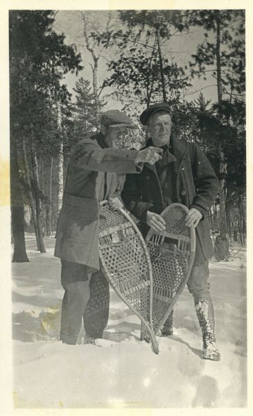 Two men are standing in the snow in the woods. Each man is holding one traditional wooden snowshoe. The man on the left has a pipe in his mouth and is pointing to the right.