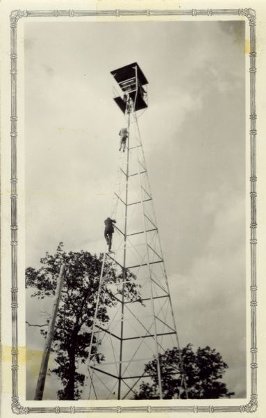View of two men on the ladder of the tower. Annotation on the back reads: "Baker tower, semi-closed, used later."