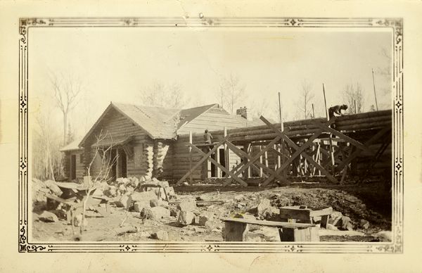 Exterior view of the building under construction, with two men working on the side of the log building on wooden scaffolds. This is probably on the site of the new Copper Falls State Park. Caption on back reads: "View of Combination Bldg. from North Facing Bad River."