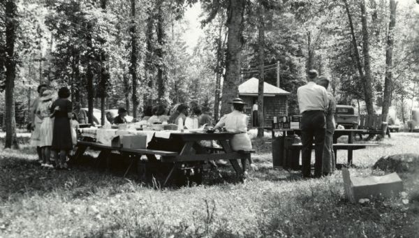 A group of people having a picnic at the Ojibwa Roadside Park. There are several cars and trucks in the background, and a small building. The picnic tables have tablecloths. The women are wearing skirts or dresses, hats and dressy shoes. The men are in shirts and slacks.