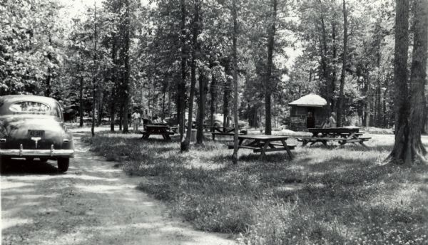 Several small groups of people are scattered about the roadside park among picnic tables. There are parked automobiles, and a small building.