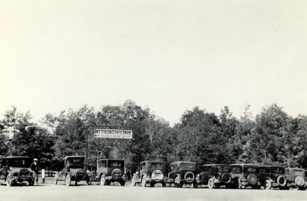 View of cars parked in the parking lot at Pattison State Park, near the entry gate and sign, which reads: "Pattison State Park, Manitou Falls — Height 165 feet.