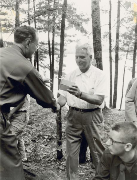 A smiling Neal Harrington is handing a box to another man. There are other people in the background and flanking Neal. They are in a state forest or park with a lake shoreline in the background.