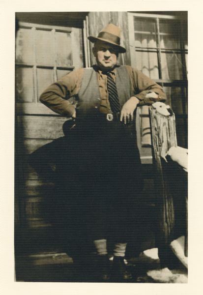 Hand-colored portrait of Pete Christensen leaning on a porch post in front of a building. He is wearing a hat slanted against the sun.