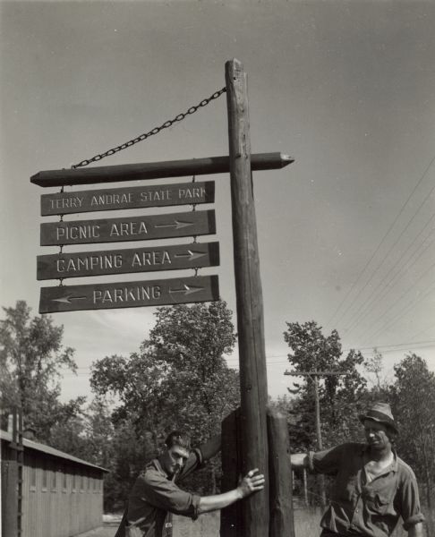 Two men are holding onto the newly installed directional sign at Terry Andrae State Park. There is a building on the left. The park was established in 1927 after the death of Mr. Terry Andrae.