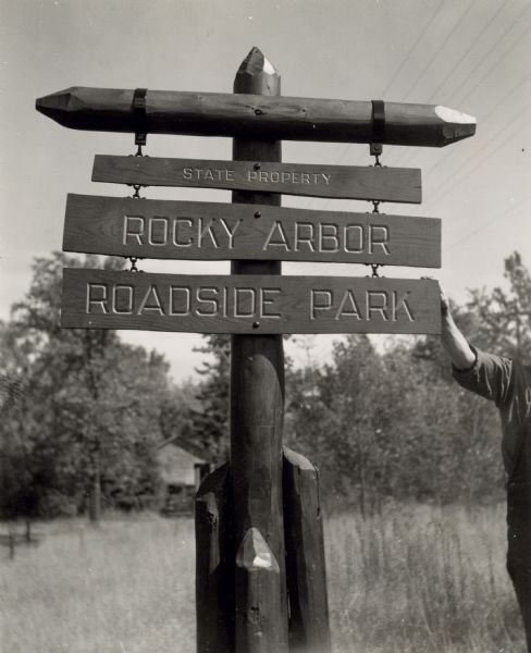 A man stands to the right holding the newly built and planted roadside park sign (only right arm visible). There is a building in the far background. Rocky Arbor Roadside Park was established in 1932 to protect sandstone outcroppings. It is now called Rocky Arbor State Park.