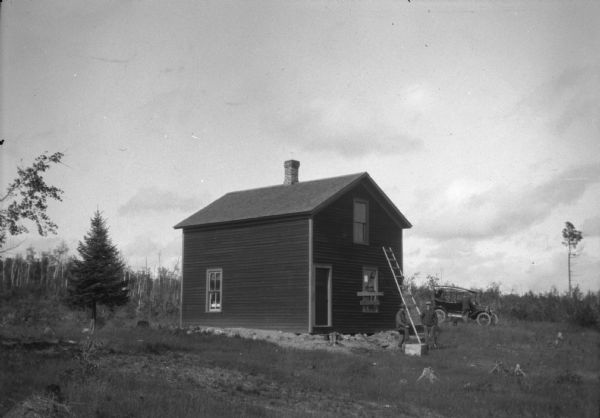 Probably a view of a ranger camp house. There is a ladder propped outside at the front, and there is a broken window with temporary board repairs. Two men are standing near the ladder. To the right of the building is an automobile. Leaning into the car is a third man. The photograph is annotated on the back: "Camp 2, no of Star Lake 1912."
