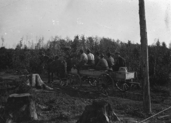 A wagon load of fire fighters is setting out or returning from the forest. The wagon has wooden wheels and is pulled by two horses. Only one of the men has turned to look at the camera, the rest of the men are facing forward.