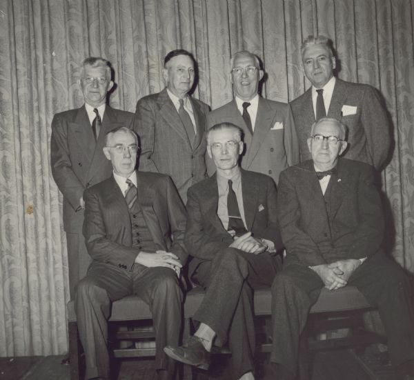 Group portrait of seven men posed in front of a curtain. C.L. (Neal) Harrington is in the back on the right. The man are attending a forestry conference.