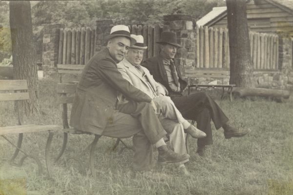Three men sitting on a park bench in the lawn outside a building which has stone columns supporting a log fence. C.L. (Neal) Harrington is on the far left of the bench.