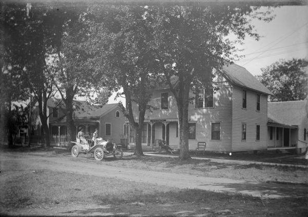 View across street of two unidentified women in dresses, and a young child, sitting and posing in an automobile parked outside of the Elm Tree Inn on E. Walter Street (later called S. 1st Avenue). In front of the Inn's front porch a man relaxes in a lawn chair with a dog lying at his feet.