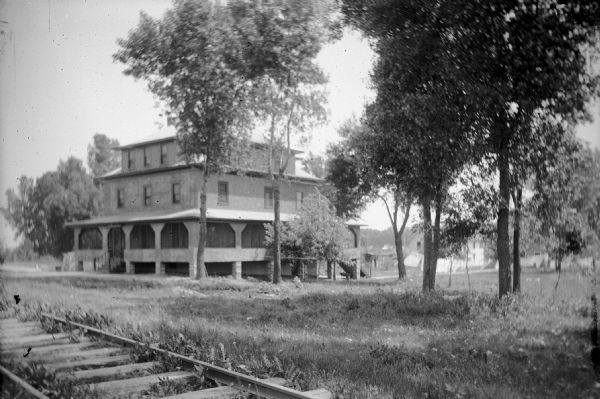 View across railroad tracks towards Bersch's Inn, a three-story house with a wrap-around porch, and flat, grassy yard. Trees flank both sides of the Inn. Located on South First Street.