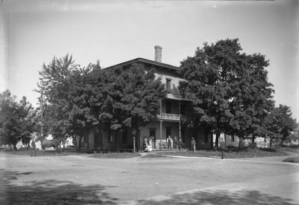 View across road of a Lake View House (1869-1950) at N. 3rd Street and N. Main Street. Three-story building with a chimney, a wrap-around balcony and a porch, obscured by trees. Under the balcony, a group of women lean against the brick wall, and a group of men stand nearby with a dog. On the far left a man stands with a horse.