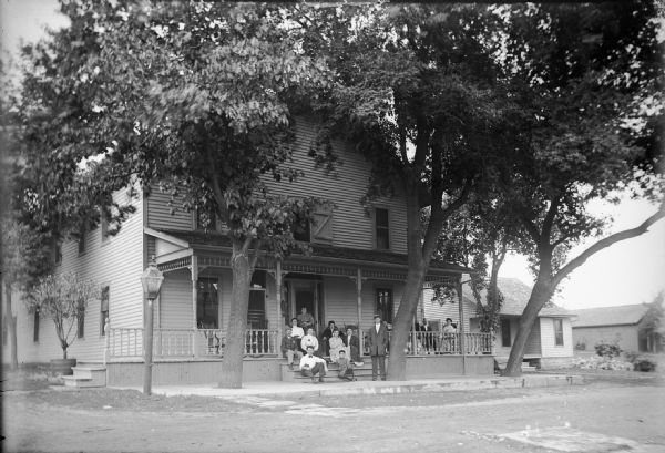View across street of a group of people posing on the front steps and and front porch of the Riverside Hotel on N. 1st Street opposite the railroad depot. Large trees shade the hotel.