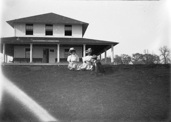 View across lawn towards group posed sitting on the lawn in front of a two-story building, the Oshkosh Power Boat Club on Lake Butte des Morts. A well-dressed man in a hat leans in to talk to two ladies wearing white dresses and Sunday hats. The back of the photograph indicates the Club was later destroyed by fire.
