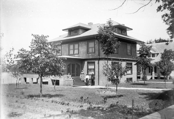 View from sidewalk of a group of unidentified people, presumably the Becker Family, sitting and standing on and near the front steps of a large house. The yard is sparely covered with young trees. In the background on the left is what may be a lake.