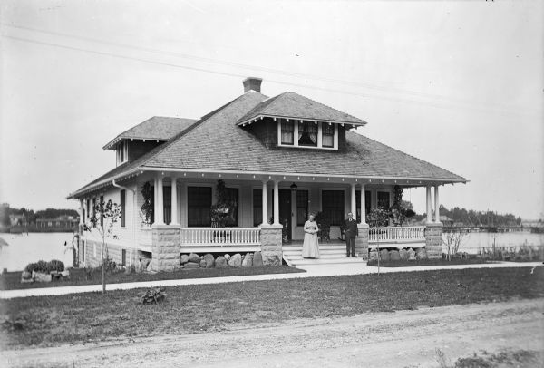 View across road towards an older man and woman standing on the front porch of a two-story house at S. 1st Avenue. The man is J.J. Phillips and the woman is unidentified. A sidewalk runs parallel to the house's foundation. A body of water (possibly Wolf River) is behind the house, and houses are along the far shoreline.