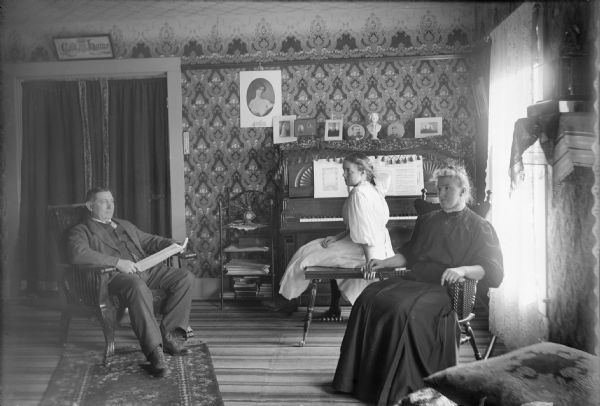 A book in his hands, Postmaster Ed King sits in his wallpapered living room, staring past his wife to the window. Edna, his daughter, is seated at the piano. She is in profile facing her father. Mrs. King sits in a rocking chair next to the window. All are staring into space. The light from the window creates a large glare. The King house was located above the Winneconne Post Office on Main Street.