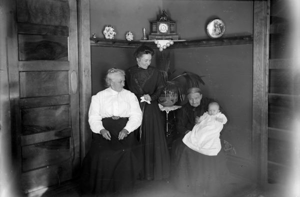 Four generations of women pose in the corner of a room. They include (left to right): Grandmother Klause, mother Ida Klause Miller, Great-Grandmother Wolf, and daughter Helen Miller (baby). A shelf holding a clock and decorative plates is on the wall behind them, and a potted plant is on a table in the corner. There are paneled doors on the left and the right.