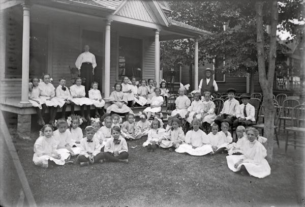 A large group of formally dressed children pose in front of the Emo house on the occasion of Cora Emo's birthday. Children sit in rows on the front porch, on the ground, and in chairs. Mrs. Emo stands on the porch behind the last row of children, and Ed Emo stands on the right behind children in the yard.