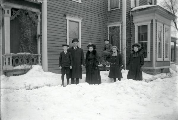 Bundled up in winter clothes, the Oakes Family poses outside the family home on the front walk. Snow covers the ground. The group includes (left to right): Hallie Oakes, Mr. and Mrs. Oakes, Grant Oakes (posed on railing behind the group), Jessie Oakes, and Irene Oakes. The house was located on S. 3rd Street. Ed Oakes was a railroad conductor. 
