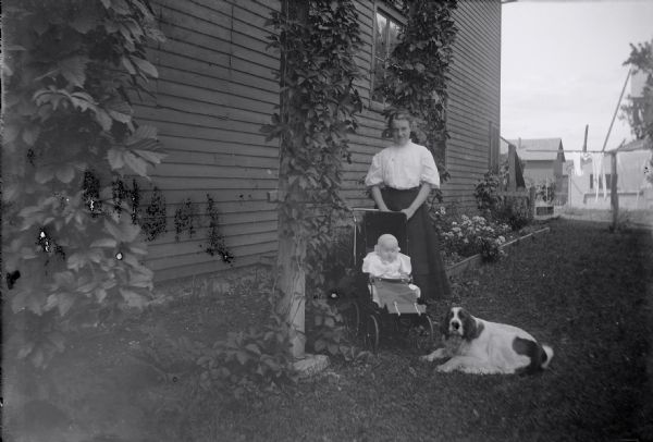 Edna King stands behind an infant in a stroller in a garden next to a wooden building. A dog is lying next to the stroller. Vines grow on lattice structures next to the building, and flowers are along the side of the house in the background.