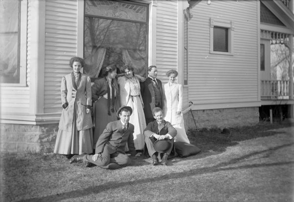 Four women and three men pose outdoors along the side of Otto Ansorge's house. They include (standing, left to right): Mrs. Ansorge, two unidentified women, George Miller, and Miss Edna Hudson, a teacher. Seated in front on the grass are, left to right: Otto Ansorge, and an unidentified man. George Miller was a local banker and Otto Ansorge was a jeweler.