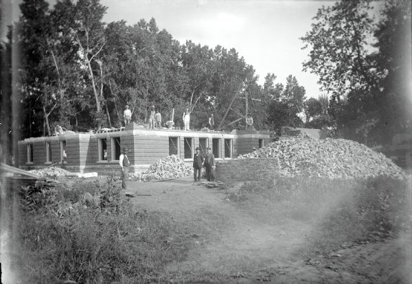 Work crew, and a young boy, posing in front and on top of the half-finished Winnebago Agricultural School. A large pile of bricks is to the right of the building site. Trees are in the background.