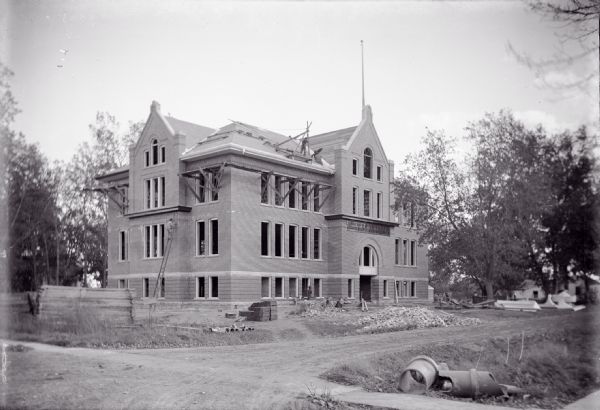 Three-quarter view of front and side of the almost-completed Winnebago School of Agriculture and Domestic Economy. Construction workers are on the building's roof, a man is climbing a ladder on the building's left side, and another man stands near the brick pile in front of the building.