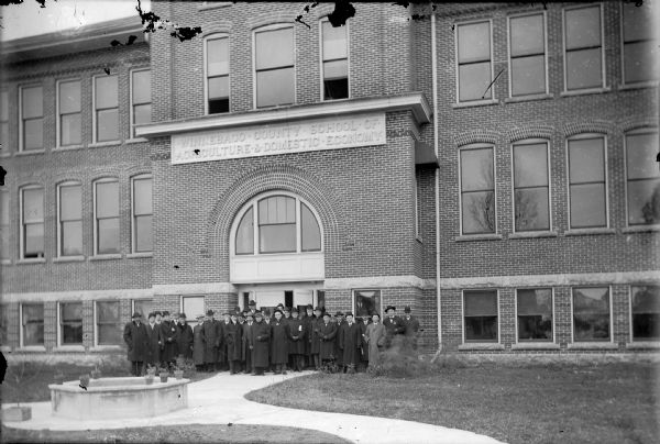 Group portrait of men dressed in coats and suits standing outside of the entrance to the Winnebago County School of Agriculture and Domestic Economy. The occasion was the first visit of Winnebago County Board members to the school.