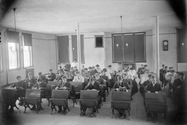 Students read attentively in one of the Winnebago County School of Agriculture and Domestic Economy's classrooms. The male students are dressed in dark suits, and the female students wear puffy-sleeved blouses and long skirts. A woman, presumably the class's teacher, stands at the back of the room.