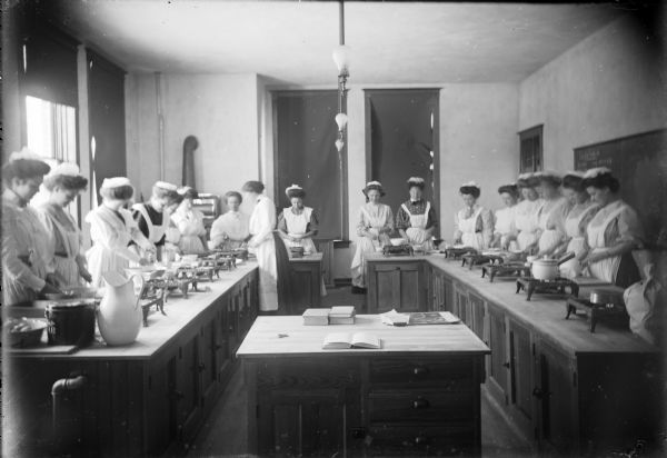 Home economics students (all women) dressed in aprons, dresses, and hats, stand in rows at counters tending pots at the Winnebago County School of Agriculture and Domestic Economy.