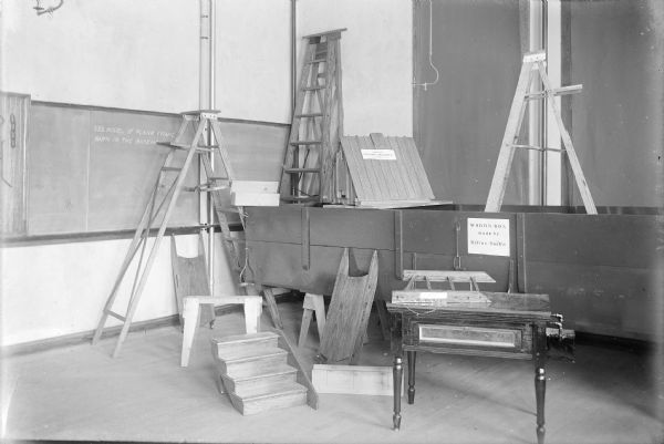 A group of ladders, stair risers, wagon box, other wooden farm equipment and furniture displayed in a classroom at the Winnebago County School of Agriculture and Domestic Economy. The objects are labeled with the names of their makers (presumably students at the school).