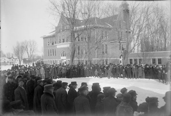 Elevated view of a large crowd of women, men and children gathered outside in the snow in front of the Winnebago County School of Agriculture and Domestic Economy. People are also looking out of the open windows of the school. Many of the men smoke pipes and cigars.