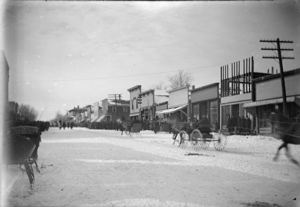 View of horse-drawn carriages and sleds racing down a snowy street lines with storefronts. A large crowd is in the distance.