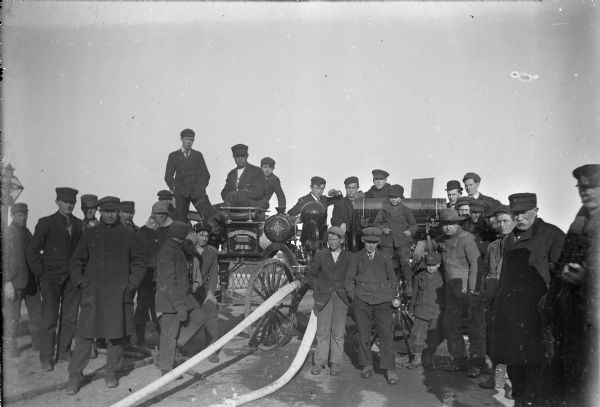 Group portrait of men and boys posing on and around a fire wagon, with a large hose attached to the front. The back of the photograph identifies the man with the moustache as Fred Klaus, but it is unclear which one of the two men on the right with a moustache is Fred.
