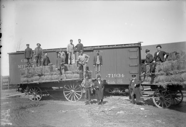 Men posing in the Winneconne Freight Yard. Some of the men stand and sit on two hay wagons, and three men stand on the ground, in front of a box car. Some of the men are dressed in work clothes, while others are wearing suits, coats and hats. A few of the men are smoking cigars.