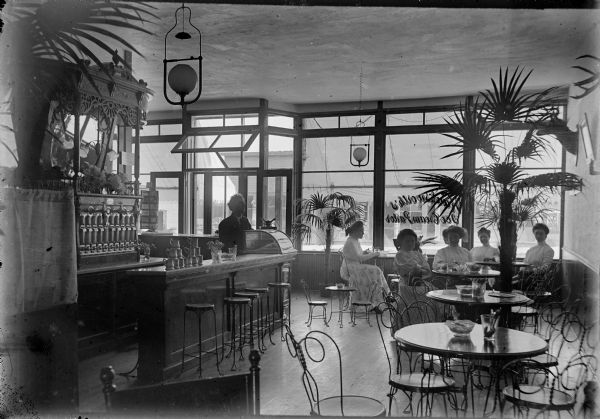 A group of five young women sit at tables in the Hawksworth Ice Cream Parlor. The woman in the center has been identified as Josephine Edith McEvoy (later Mrs. E.C. Oakes). Bright light from the large plate glass windows and front door behind the women creates dramatic shadows within the parlor. Ferns and palm trees decorate the room. Calvin Hawksworth, the establishment's owner, looks on from behind the counter. A large soda fountain with a mirror is behind him. A child-size set of two chairs and a table are just in front of the counter.