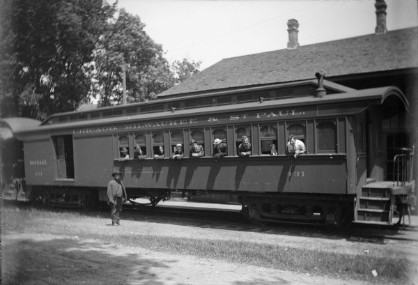 Eight men and one dog lean their heads outside of the windows of a passenger car caboose of the Chicago, Milwaukee, and St. Paul railway for a group portrait. One man stands on the ground in front of the train car. The location is presumably the Winneconne Railroad Station.