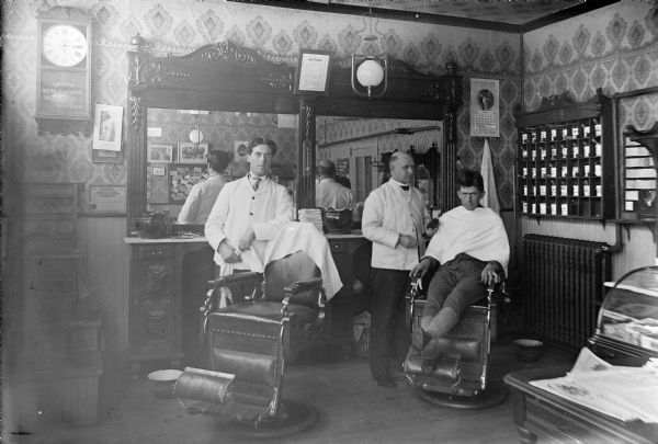 Two barbers: Elmer Lee (left) and William Goold (right), stand beside their barber's chairs while an unknown male patron sits in one of the chairs. Reflections of the men are in the large mirrors behind them on the wall.