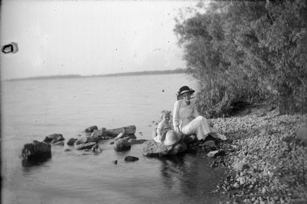 Wearing a long, white dress, white shoes and a large, ribboned hat, Mary Ansorge poses on a large stone on the rocky bank of Lake Winneconne. Her daughter, Betty, sits on a stone beside her, smiling and holding a large straw hat on her knees. Dense foliage is on the right. The distant shore of the other side of Lake Winneconne is in the background.