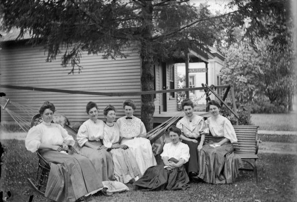 Seven women posed in yard. A woman, Ida Lund, sits in a rocking chair on the left, three more, Maude Riley; Maggie Riley; Lydia Mader, pose sitting in a hammock, one woman, Nona Riley, sits on the ground, and on the right, Lulu Lee and Ida Klaus sit on a bench. Two houses are in the background, and the one directly behind the group has a signon the porch that reads: "?R. Hughes."