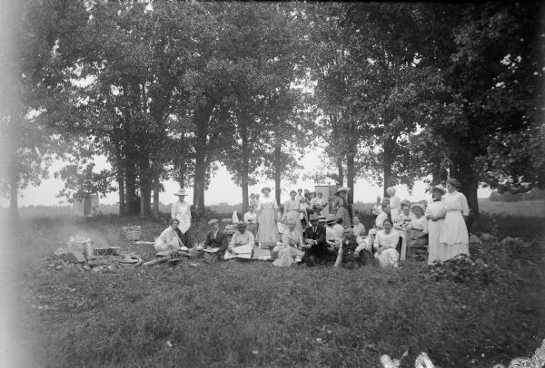 Group portrait of a large group of people posing sitting and standing amongst the trees at Larsley's Point at Lake Winneconne. Some individuals pose purposefully while others appear to be unaware that a portrait is being taken. Most of the women wear light-colored dresses, and a number of the men are wearing suits. Some in the group are posed on the ground on cloths set with picnic baskets, and another group is posed sitting around a table.