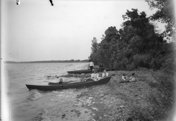 View along shoreline towards two young men and six young children posing on the rocky beach on Lake Winneconne. Four of the children sit in a kayak pulled up on the shore, while two others play on the rocky shoreline nearby. In the background the two young men pose next to a group of canoes also pulled up on the shoreline. Dense foliage and trees are along the shoreline.