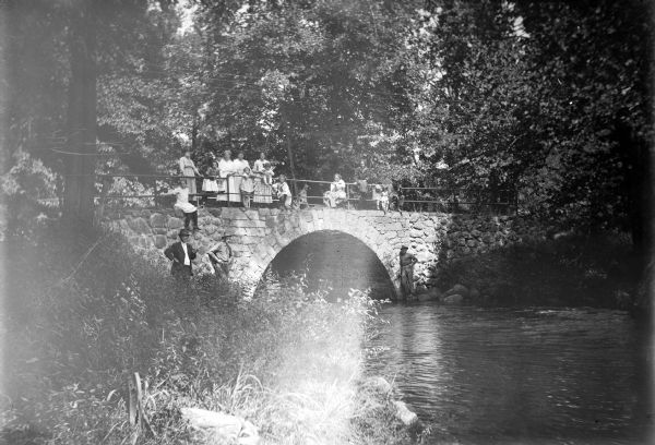 View from shoreline of a group of children and adults posing on and around a stone bridge over the river. Women and children stand and sit on the bridge's metal railing. Three men pose on the left and right banks against the stone foundation arch of the bridge. The back of the photograph identifies the area as Pine River Camp Ground.